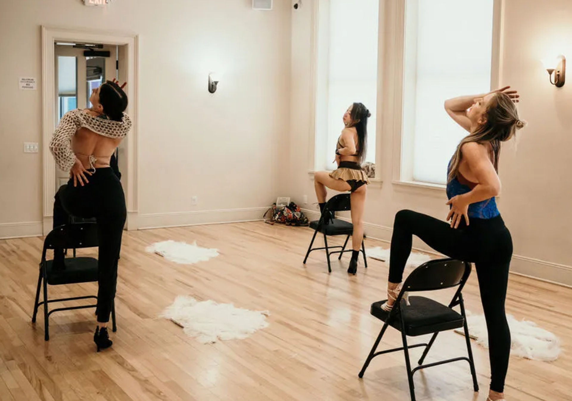 Three women in dancewear striking choreographed chair-dance poses in a bright hardwood dance studio with folding chairs and white faux-fur floor accents — energetic chair dance/fitness class