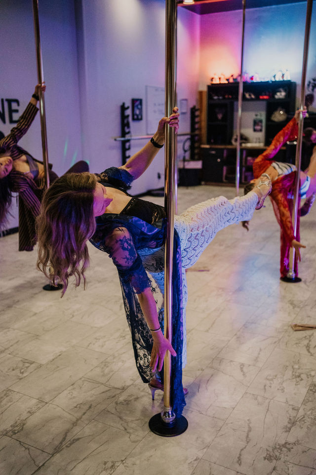 Women practicing pole fitness in a colorful dance studio, leaning sideways on chrome poles in lace outfits and heels over a marble floor with purple-pink ambient lighting.