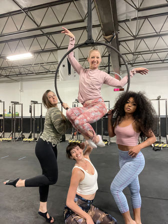 Four women in an indoor aerial fitness studio pose on a suspended hoop (lyra), group workout in colorful activewear inside a warehouse-style gym