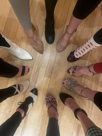 Top-down circle of women's shoes on a hardwood floor — mixed styles and colors including cowboy boot, ankle-strap heels, ballet flats, two-tone oxfords, strappy sandals and patterned socks, playful group shoe selfie.