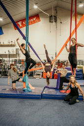 Group of women practicing aerial silks in an indoor gym — colorful fabric hammocks and padded mats with one woman suspended upside down while others climb and assist in an aerial yoga/acrobatic class.