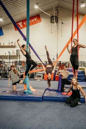 Group of women practicing aerial silks in an indoor gym — colorful fabric hammocks and padded mats with one woman suspended upside down while others climb and assist in an aerial yoga/acrobatic class.