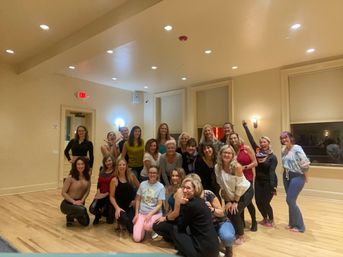 Group of about 20 women smiling and posing on a wood floor in a bright indoor studio or community room with recessed lights and windows — post-class photo from a dance/fitness/yoga workshop.
