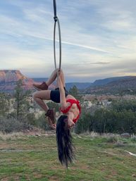 Aerial hoop artist hanging upside down from a suspended hoop, wearing a red top and boots against a red rock canyon backdrop and pastel sunset sky over desert greenery.