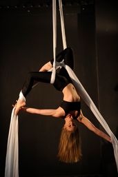 Gravity-defying aerialist in black athletic wear suspended upside-down on white silks in a dark studio, holding her foot in a graceful backbend.