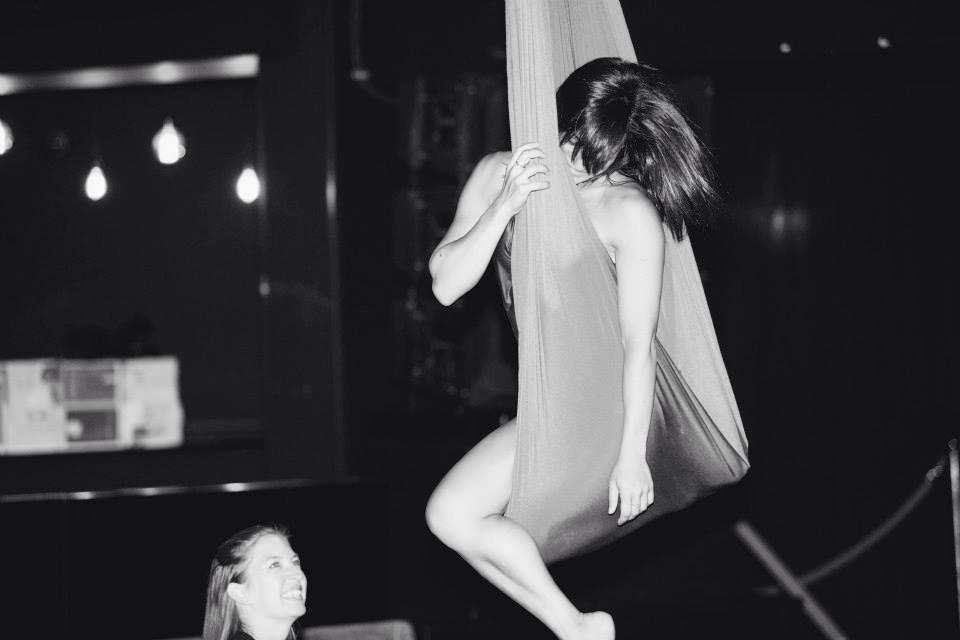 Black-and-white photo of a female aerialist suspended in an aerial silk hammock in a dimly lit indoor studio while a smiling spotter watches below