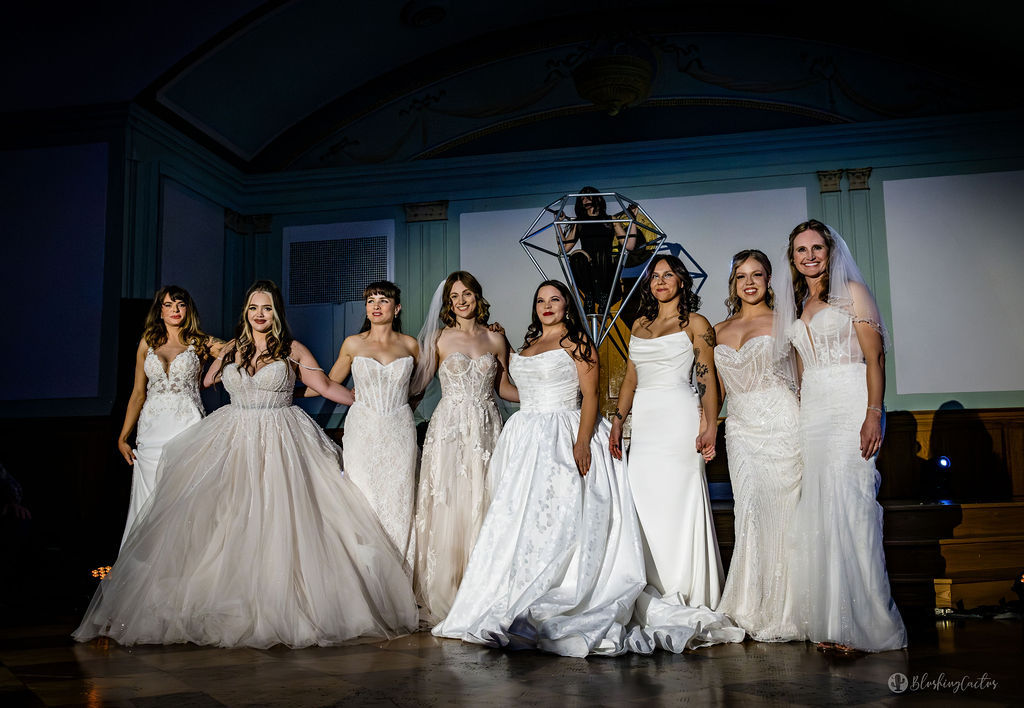Bridal fashion show: eight women in varied wedding gowns (ball gown, A-line, mermaid, sheath, lace) standing on a grand indoor ballroom runway under warm stage lighting, smiling and holding hands.