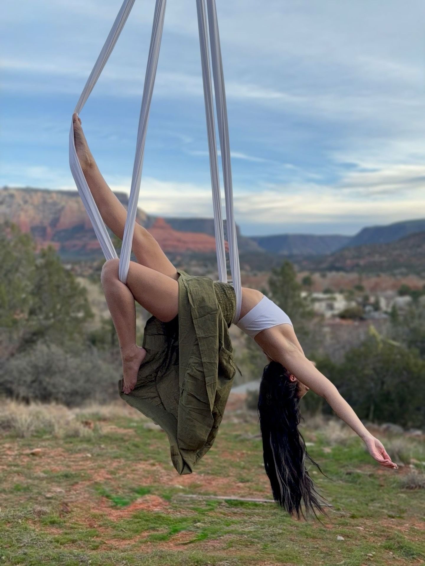 Person doing aerial silk yoga suspended upside-down from white silks above a red‑rock desert canyon, wearing a white top and olive skirt with long hair trailing against a blue sky.