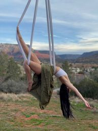 Person doing aerial silk yoga suspended upside-down from white silks above a red‑rock desert canyon, wearing a white top and olive skirt with long hair trailing against a blue sky.