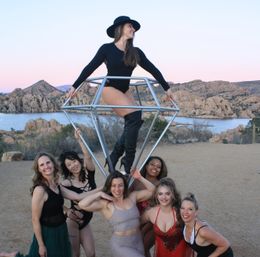 Seven women in performance costumes pose outdoors at a lakeside sunset with granite rock formations; one woman in a black bodysuit and hat stands inside a geometric metal diamond prop while the others smile below.