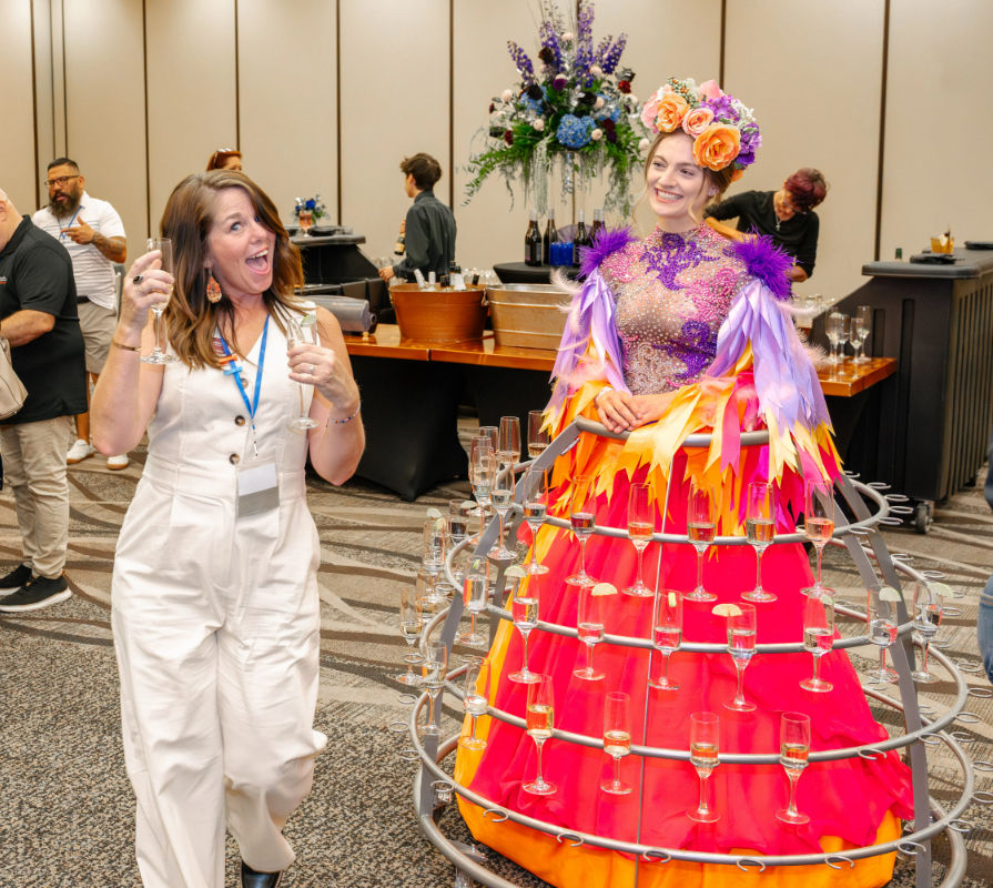 Woman in a white jumpsuit toasting with champagne beside a smiling performer in a bright floral headpiece and multi-tiered skirt holding dozens of champagne flutes at an indoor conference cocktail reception.