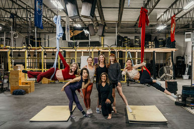 Group of eight women in a warehouse-style fitness studio posing for a group fitness photo, two suspended on red and blue aerial silks while others stand and kneel on mats amid gym equipment and weight stacks.