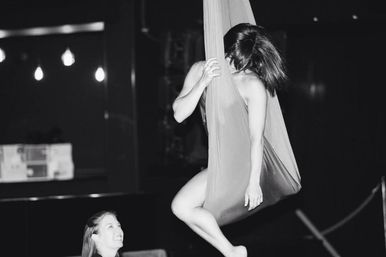 Black-and-white photo of a female aerialist suspended in an aerial silk hammock in a dimly lit indoor studio while a smiling spotter watches below