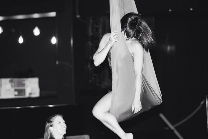 Black-and-white photo of a female aerialist suspended in an aerial silk hammock in a dimly lit indoor studio while a smiling spotter watches below