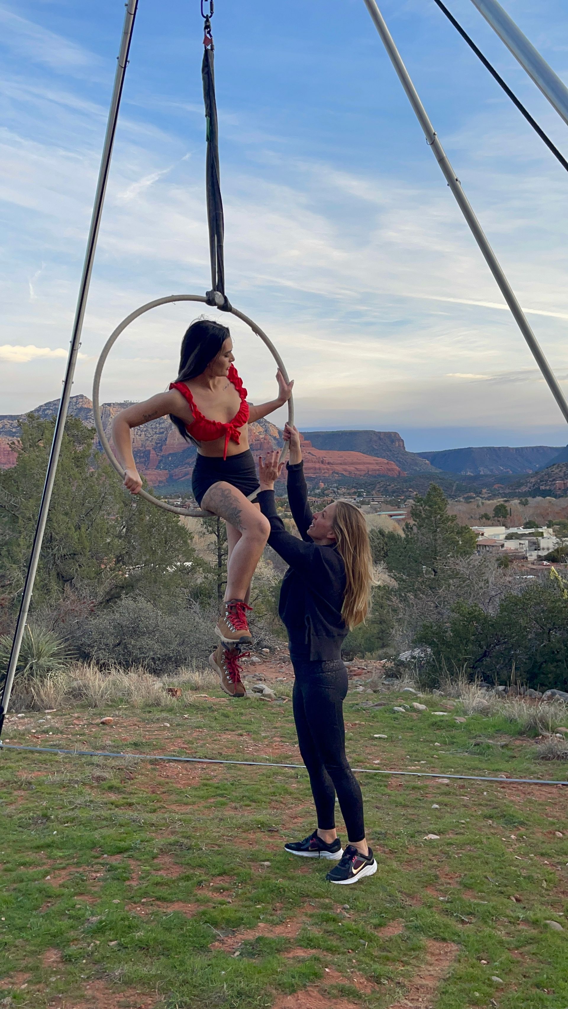 Outdoor aerial hoop (lyra) practice over red rock canyon at dusk — performer in a red crop top on a suspended hoop while an instructor spots from a grassy desert overlook.