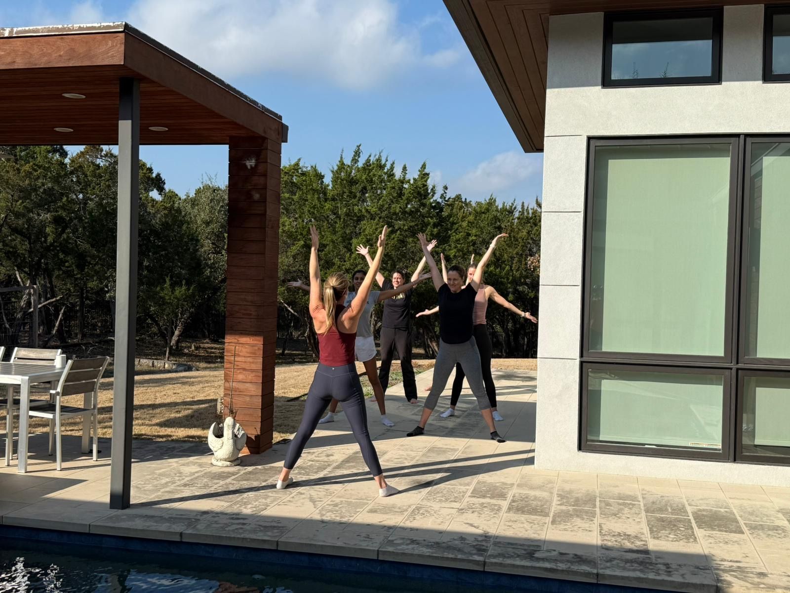 Five people doing a group workout (jumping jacks) on a modern home's poolside patio in a sunny backyard with trees