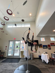 Aerial hoop performer suspended upside down on a freestanding pole hoop in a bright modern lobby or event space, wearing a black-and-gold sequined costume and heels, playfully holding a bottle beneath hanging circular mobile and near small reception tables.