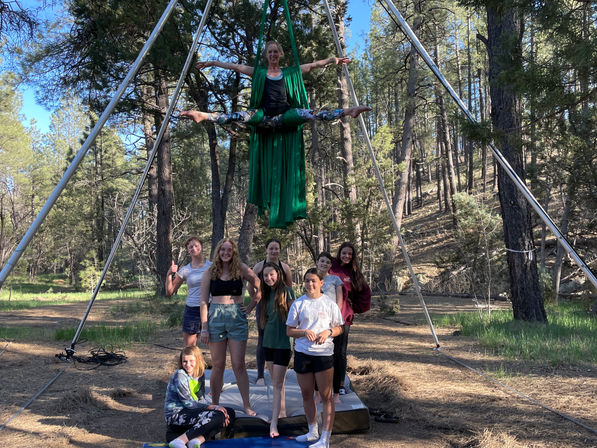 Aerial silks performer in bright green fabric holding a high straddle under a metal rig in a sunlit pine forest, with eight young people posing on crash mats beneath — outdoor aerial silks workshop in a wooded clearing.