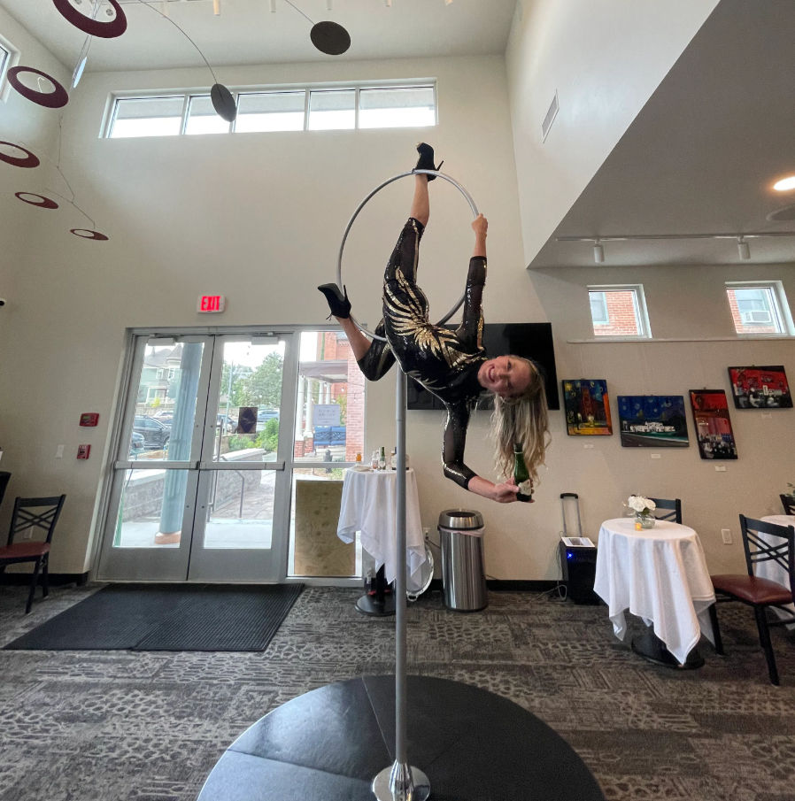Smiling aerialist in a black sequined costume performs upside-down on a freestanding aerial hoop (lyra) in a bright indoor event space by glass double doors, holding a bottle with tables and artwork in the background.