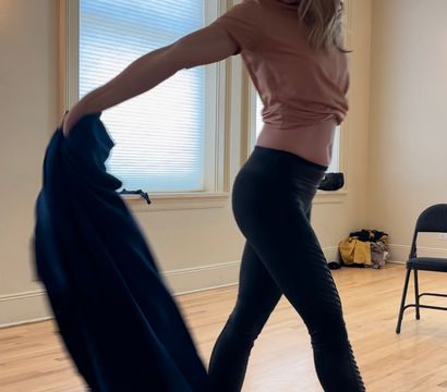 Woman striking a dramatic dance pose in a sunlit dance studio on hardwood floors, wearing a peach top, black leggings and strappy heels while swinging a flowing navy fabric.