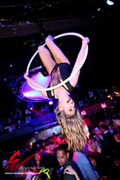 Upside-down aerial hoop acrobat suspended above a lively nightclub crowd, blonde performer smiling amid colorful stage lights and club atmosphere