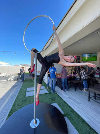 Smiling aerial hoop acrobat in black outfit and red heels posing on a pole-mounted hoop at a sunny backyard patio party with guests in the background.