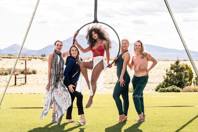 Five women pose in a sunny outdoor group portrait on a grassy field with desert mountains in the background; a central aerialist in a red lace top and flowing white skirt sits in a suspended aerial hoop (lyra) while four women stand smiling around her.