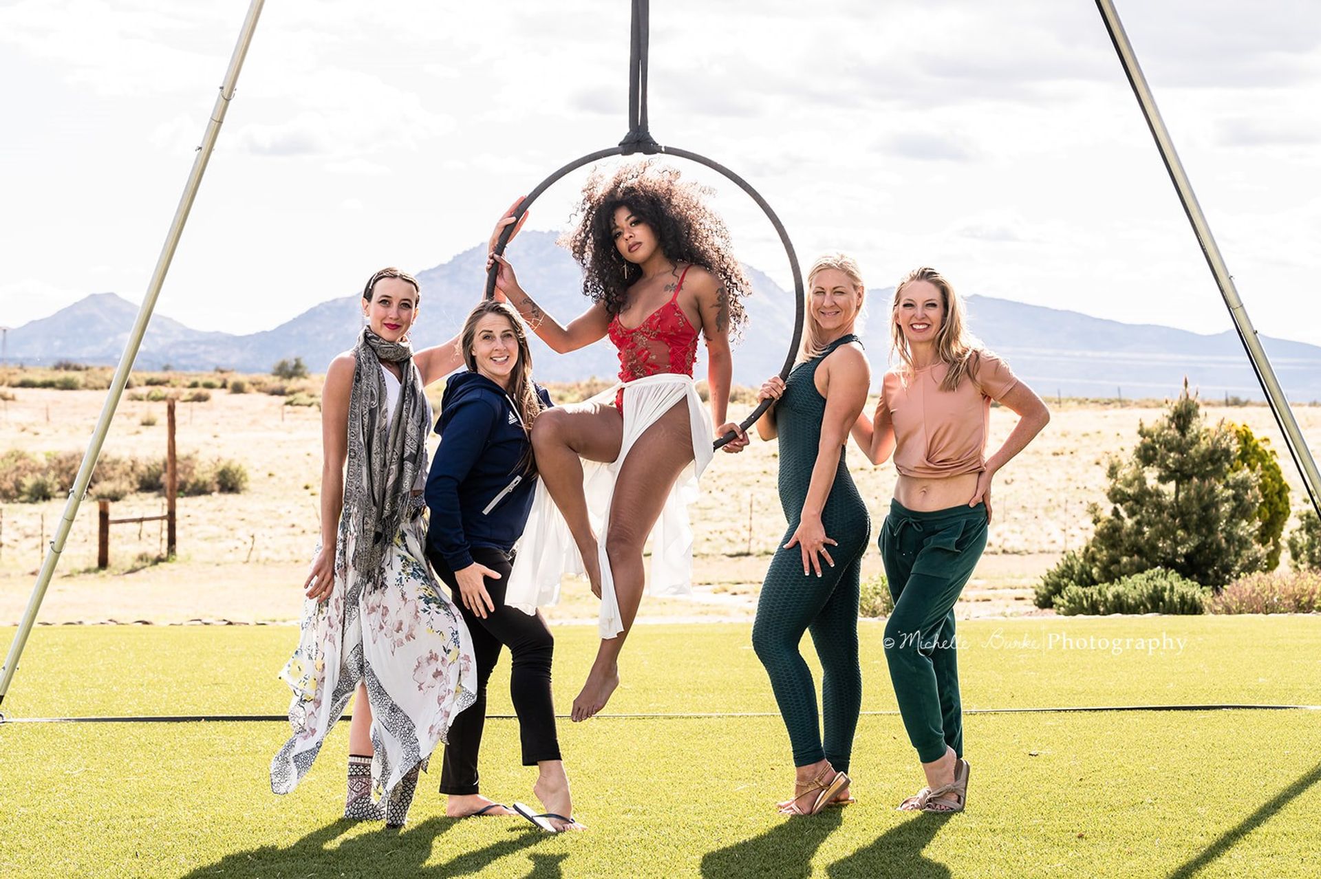 Five women pose in a sunny outdoor group portrait on a grassy field with desert mountains in the background; a central aerialist in a red lace top and flowing white skirt sits in a suspended aerial hoop (lyra) while four women stand smiling around her.