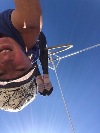 Person inverted on an outdoor aerial hoop (lyra) against a clear blue sky, wearing a patterned cap, leggings and pink-striped toe socks — sunny aerial fitness scene.