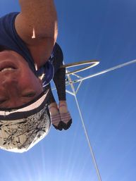 Person inverted on an outdoor aerial hoop (lyra) against a clear blue sky, wearing a patterned cap, leggings and pink-striped toe socks — sunny aerial fitness scene.