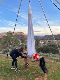 Participant reclined in a white aerial silk hammock while an instructor adjusts the fabric, set against Sedona red rock mesas and a blue sky sunset backdrop.