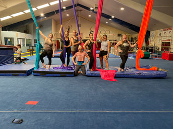 Seven women in an aerial silks class pose with teal, purple, pink and orange silks on padded mats inside an indoor gymnastics gym, surrounded by bars, rings and training equipment.