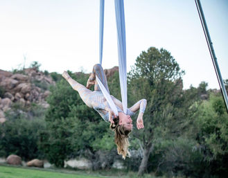 Aerial silk performer suspended upside down from white silks, wearing a sparkly bodysuit above a grassy area with rocky, tree-lined landscape