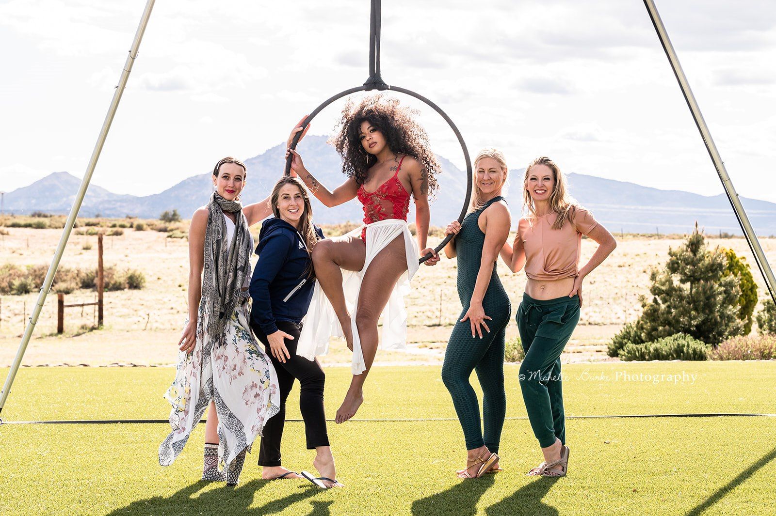 Five women pose outdoors around a suspended aerial hoop on green turf with desert mountains behind them; the central performer in a red lace top and white skirt sits in the hoop while four women smile and support her in a playful photoshoot.