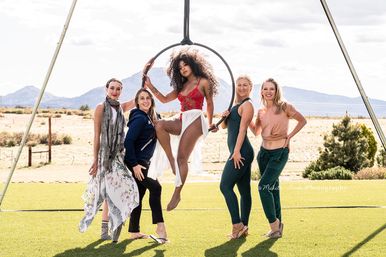 Five women pose outdoors around a suspended aerial hoop on green turf with desert mountains behind them; the central performer in a red lace top and white skirt sits in the hoop while four women smile and support her in a playful photoshoot.