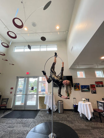 Aerial acrobat in a black sequined costume performs upside-down on a silver aerial hoop in a bright indoor event space or lobby, smiling and holding a bottle near glass entrance doors, with a modern hanging mobile and small cocktail tables with white tablecloths.