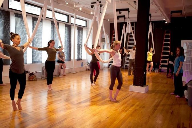 Group aerial yoga class in a bright hardwood studio, participants barefoot practicing stretches using white aerial silks suspended from the ceiling while an instructor observes.