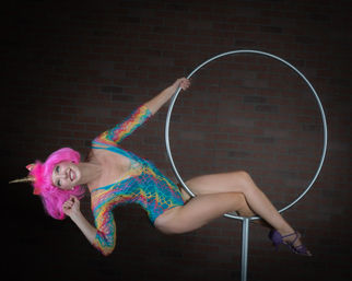 Aerial hoop performer in a rainbow-patterned bodysuit and bright pink wig with a gold unicorn horn, posing sideways on a metal lyra against a brick wall, wearing purple heels.