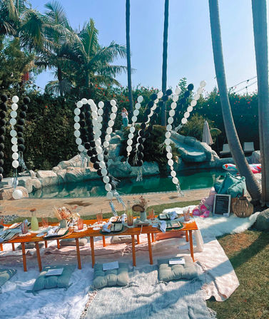 Sunny backyard pool picnic under palm trees with black-and-white balloon garlands, low wooden table, floor cushions and a rock slide next to an inflatable float.