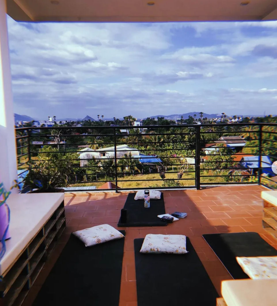 Rooftop terrace with yoga mats and floral cushions on terracotta tiles, black railing framing a tropical landscape of palm trees, scattered houses and distant mountains under a blue cloudy sky.