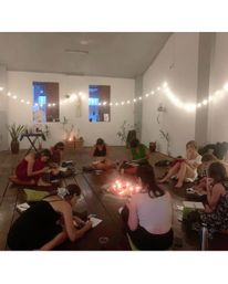 Cozy candlelit studio circle of people seated on a wooden floor, writing in notebooks under string lights and houseplants during a relaxed journaling/meditation workshop.