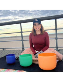 Smiling woman wearing a baseball cap, seated on a beach pier at sunrise, wrapped in a blanket with blue, green, and orange crystal singing bowls arranged on a mat in front of her.