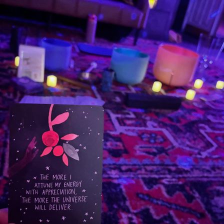 Affirmation card in foreground reading "The more I attune my energy with appreciation, the more the universe will deliver," set on a colorful patterned rug with crystal singing bowls, flickering LED candles and purple-blue mood lighting — cozy indoor meditation/sound-bath scene.