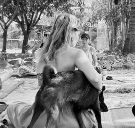 Back view of a woman sitting on a mat in a shaded outdoor garden, cuddling a small dark dog while others relax nearby