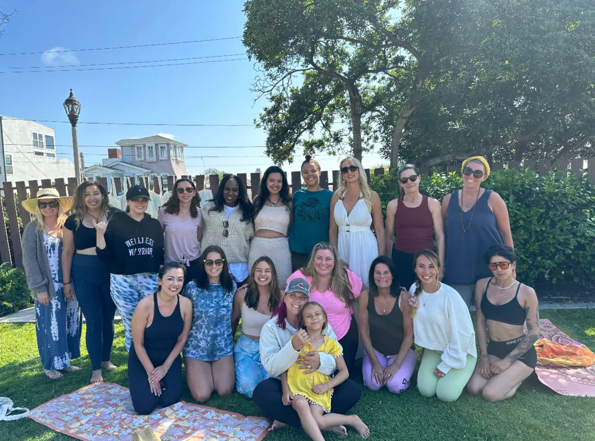 Smiling group of women and a child gathered on a sunlit backyard lawn, posing in casual and athletic outfits in front of a wooden fence with trees and residential beach-style houses in the background — cheerful outdoor group photo.