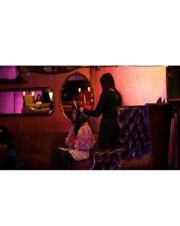 Moody urban lounge with pink-purple neon, one person holding a giant wine glass over a seated friend on a sofa, ornate mirrors and a tufted leather armchair