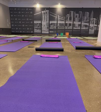 Indoor yoga studio setup with rows of purple mats and black blocks on a polished floor, front wall featuring a monochrome bridge and city skyline mural.