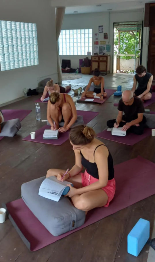 Participants in a bright indoor yoga studio sitting on mats and bolsters, writing in journals during a mindfulness workshop — wooden floors, natural light and yoga blocks visible.