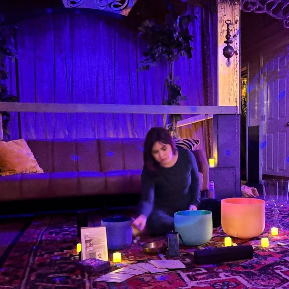 Person seated on a patterned rug in a purple-lit indoor meditation space, playing colorful crystal singing bowls surrounded by small LED candles, cards and cozy sofa in the background.
