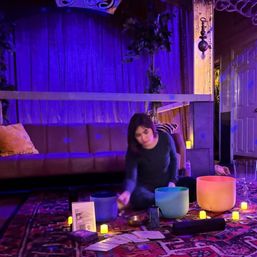 Person seated on a patterned rug in a purple-lit indoor meditation space, playing colorful crystal singing bowls surrounded by small LED candles, cards and cozy sofa in the background.